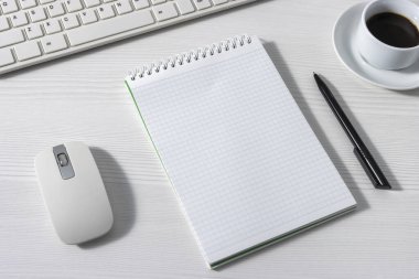 Fare, klavye ve beyaz ofis masasında beyaz bir kahve fincanının yanında boş bir not defteri.