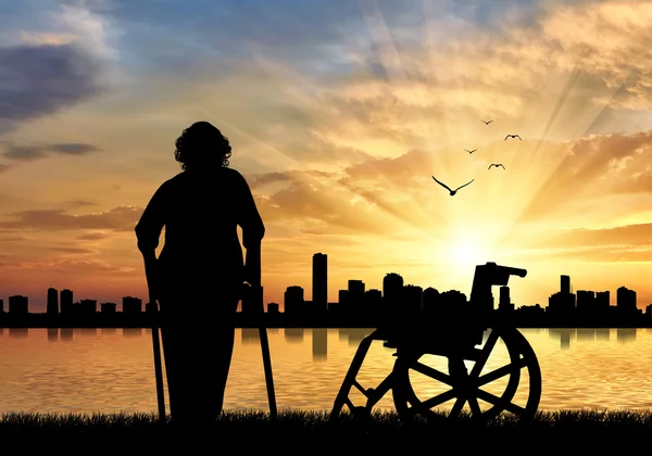 Silhuetten av en gammal kvinna på kryckor på en bakgrund av havet solnedgång urban och rullstol — Stockfoto