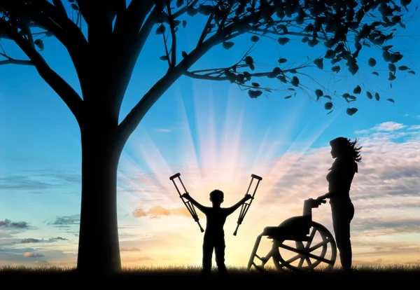 Happy disabled Child is invalid with crutches beside wheelchair and nurse near tree