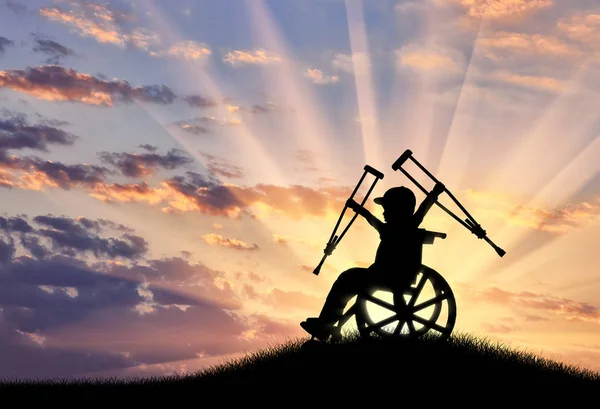 Menino feliz sentado em cadeira de rodas com muletas por do sol — Fotografia de Stock