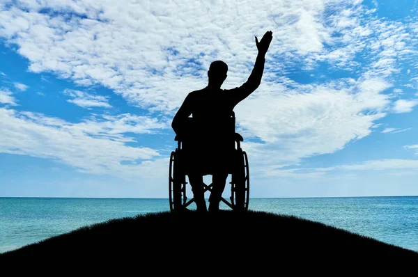 Silueta de un hombre discapacitado en silla de ruedas agitando su mano sobre una colina cerca del mar — Foto de Stock