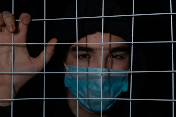 a teenager with black eyebrows in a black hood and an individual mask stands behind bars, his hand on the bars. Focus on the grill. Syrian refugee problem in Europe