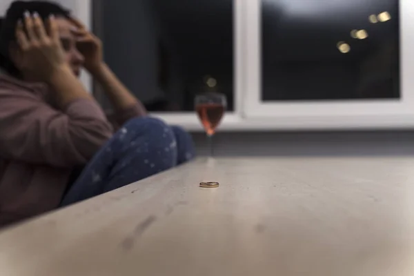 woman is sitting at the table and crying. in front of her is a bottle of wine. the wedding ring lies in front of her on the table. topic of depression and alcoholism in connection with a divorce.