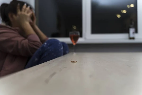 woman is sitting at the table and crying. in front of her is a bottle of wine. the wedding ring lies in front of her on the table. topic of depression and alcoholism in connection with a divorce.
