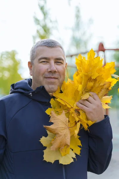 portrait of a gray-haired man in a blue jacket with an armful of autumn leaves