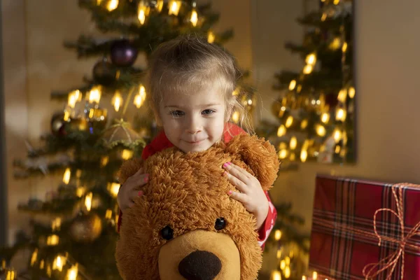 a girl of three years in a red dress hugs a big brown bear near the Christmas tree. Happy smile. Blurred background. New Year holidays concept.