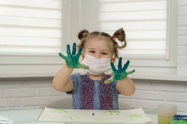 Little girl in mask paints with fingers.girl joyfully raised her hands in raska up, self-isolation, coronavirus covid-19, stay home