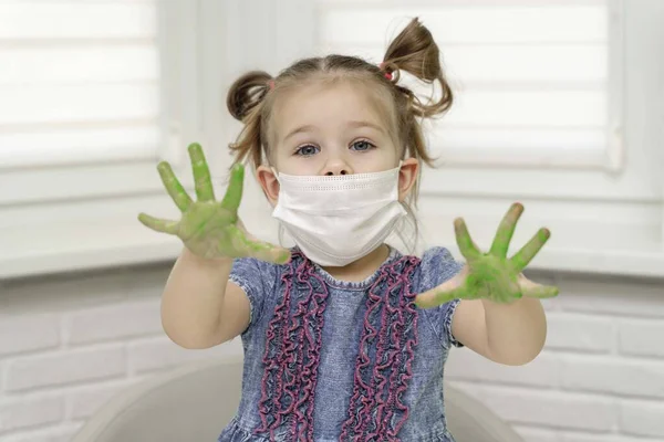 Little girl in mask paints with fingers.girl shows us her green palms in paint, self-isolation, coronavirus covid-19, stay home