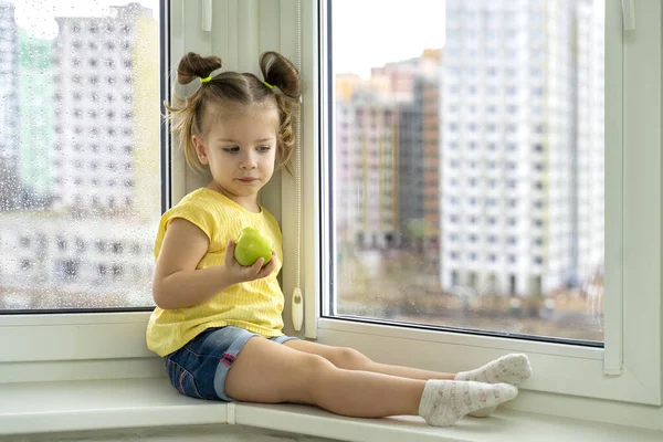 a little girl sits by the window, sad and holds an apple in her hands. quarantined children