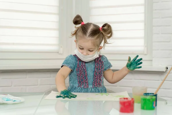 Little girl in mask paints with fingers.girl makes a print on paper with her right green palm in the paint, self-isolation, coronavirus covid-19, stay home
