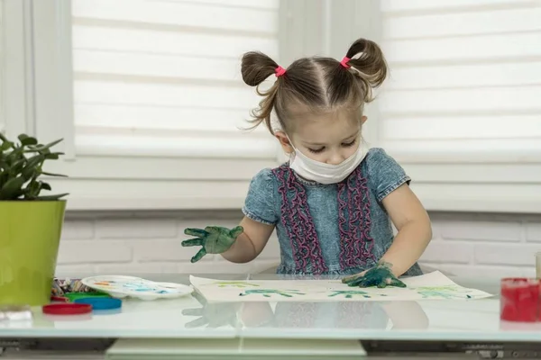 Little girl in mask paints with fingers.girl makes a print on paper with her left green palm in the paint, self-isolation, coronavirus covid-19, stay home