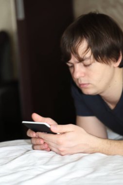 Koyu renk saçlı bir adam sabahları elinde telefon ve e-kitapla yatakta uzanıyor. Sabahları aletlere bağımlı oluyor.