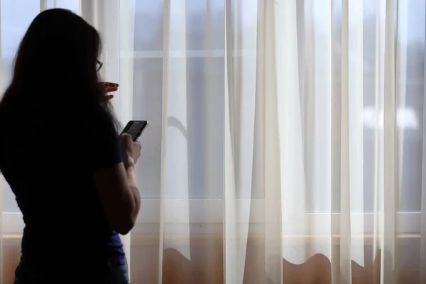 A menina com o telefone está de pé na janela e chorando Imagens De Bancos De Imagens