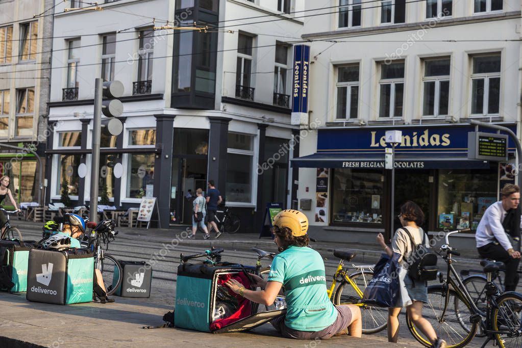 Ghent, Belgium - August 28, 2017: Worker of Deliveroo resting with people walking and cycling in the old town of the medieval city of Ghent, Belgium