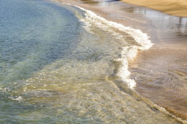 Güneşin altında kumlu sahilde küçük dalgalar kopuyor.