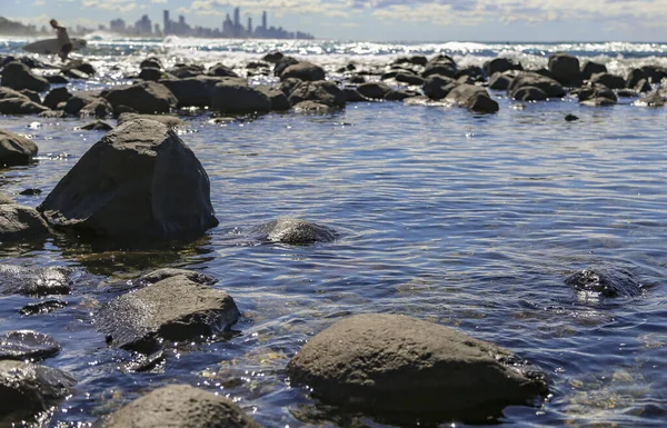 Sörfçünün bulanık arka planına sahip küçük sakin bir kaya havuzu. Sörf tahtasıyla okyanus ve kayaların arkasındaki yüksek binalarla birlikte suyun dışında yürüyor.