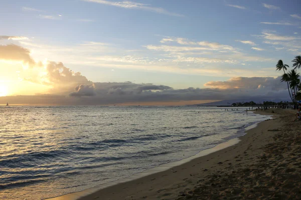 Kumlu bir kumsalda gün batımı, küçük dalgalı palmiye ağaçları ve kum, ufukta alçak bulutlar parlak güneşin filtrelediği ve uzaktaki tek yelkenli..