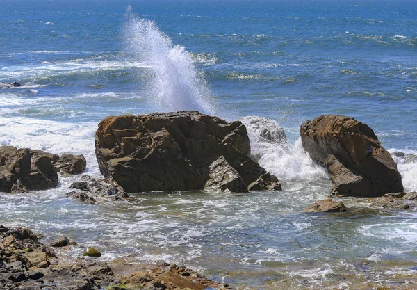 Bir dalga bir kayanın üzerine çarpıyor. Uzakta parlak mavi okyanusla havaya püskürtüyor.