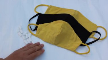 Siyah ve sarı tıbbi maske ve elle sayım hapları. Koronavirüsten korunma. Bir kadın beyaz arka planda beyaz bir maskenin yanında beyaz haplar sayıyor. Yakın plan.