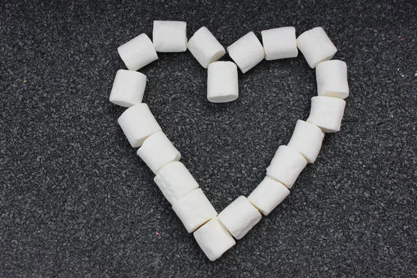 Marshmallows laid out in the shape of a heart isolated on a gray stone background. Bunch of small white marshmallows laid out in the form of a heart on a table. Sweets on a close-up.