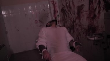 Deli gömleği giymiş deli bir adam sandalyeye bağlanmış, duvarları kanlı bir odada aşırı doz almış katil bir psikopat.