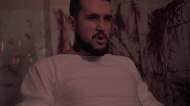 Gekke man in dwangbuis vastgebonden aan een stoel, moordende psychopaat in een kamer met bebloede muren. — Stockvideo