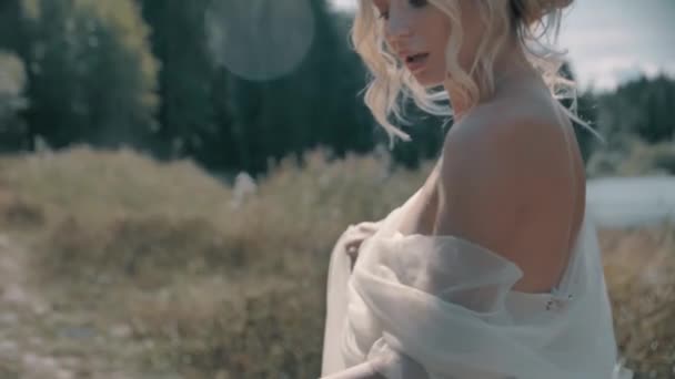 Blond meisje in een witte jurk zitten in de buurt van het meer — Stockvideo