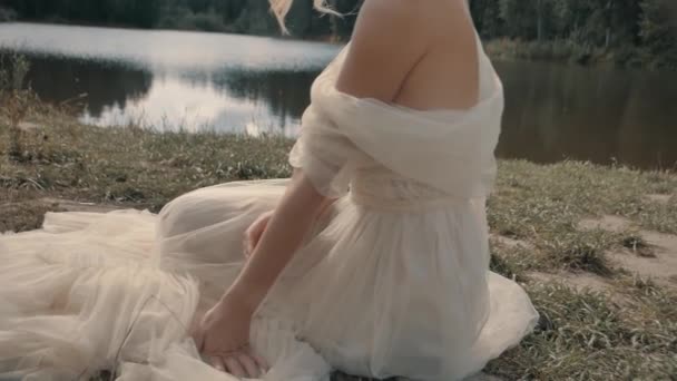 Блондинка в белом платье сидит у озера крупным планом — стоковое видео