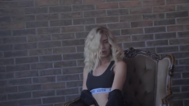 Gyönyörű fiatal szőke fekete fehérneműben meztelenül ül egy székben téglafal — Stock videók
