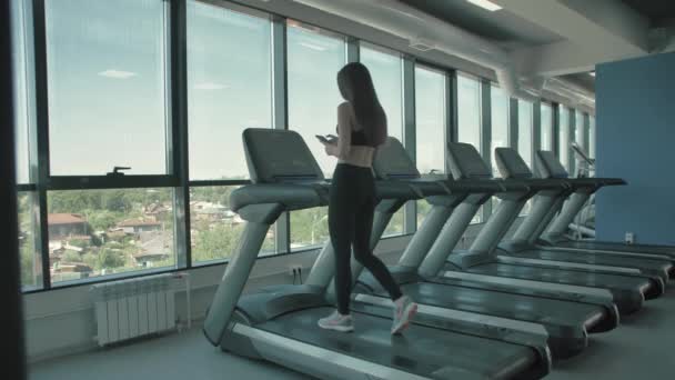 Hermosa chica deportiva escuchar música desde el teléfono inteligente con aplicación que se ejecuta en la cinta de correr — Vídeo de stock