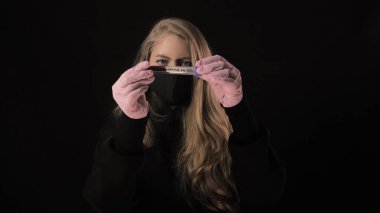 Siyah maskeli kız üzerinde kan olan bir test tüpü ve Covid-19 yazısını tutuyor. Siyah arka planda izole edilmiş. Sağlık ve tıbbi konsept. Koronavirüs salgını