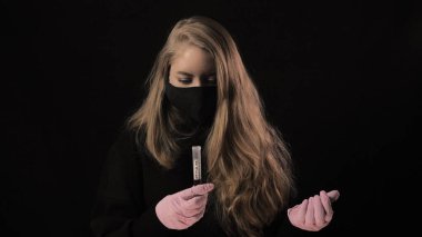 Siyah maskeli kız üzerinde kan olan bir test tüpü ve Covid-19 yazısını tutuyor. Siyah arka planda izole edilmiş. Sağlık ve tıbbi konsept. Koronavirüs salgını