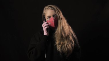 Beyaz kurdeleli ve kırmızı maskeli Covid 19 yazılı kız akıllı telefondan selfie çekiyor. Siyah arka planda izole edilmiş. Sağlık ve tıbbi konsept. Koronavirüs salgını