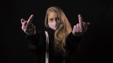 Girl in a pink mask and a ribbon with the inscription covid 19 show gesture Fuck. Isolated on black background. Health care and medical concept. Close up portrait . 4k. Coronavirus Epidemic