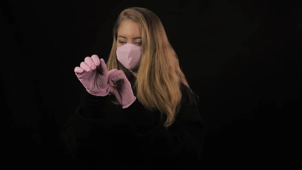 Chica de color rosa máscara médica endereza guantes de color rosa en la mano. Aislado sobre fondo negro. Salud y concepto médico. Cierre el retrato. 4k. Coronavirus Epidemia, enfermedad, pandemia —  Fotos de Stock