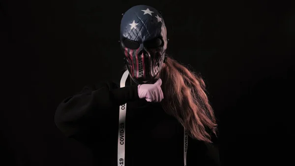 Menina em uma máscara de cor assustadora EUA e uma fita branca com a inscrição covid 19 mostra um gesto de morte. Isolado em fundo preto. Cuidados de saúde e conceito médico. Epidemia por Coronavírus — Fotografia de Stock