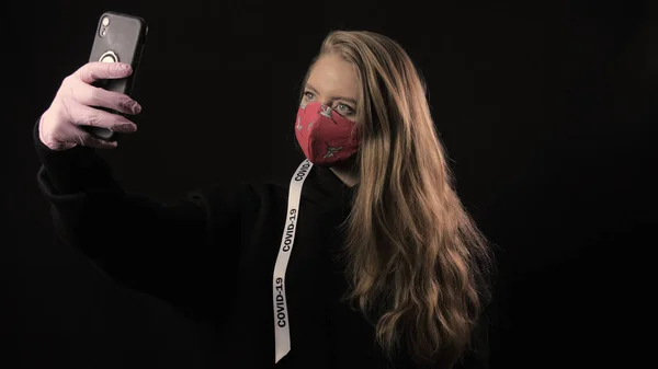 Chica con una cinta blanca y la inscripción covid 19 en máscara roja toma selfie en el teléfono inteligente. Aislado sobre fondo negro. Salud y concepto médico. Epidemia del Coronavirus — Foto de Stock