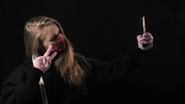 Chica con una cinta blanca y la inscripción covid 19 en máscara roja toma selfie en el teléfono inteligente. Aislado sobre fondo negro. Salud y concepto médico. Epidemia del Coronavirus — Foto de Stock