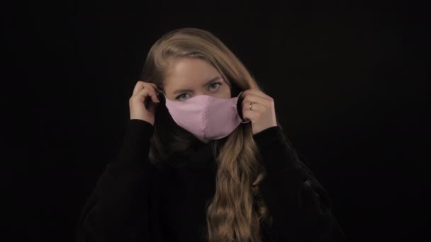 Mladá žena se zlatými vlasy si nasadí růžovou lékařskou masku. Izolované na černém pozadí. Zdravotní péče a lékařská koncepce. Zavřít portrét. 4k. Koronavirus Epidemie, onemocnění, pandemie — Stock video