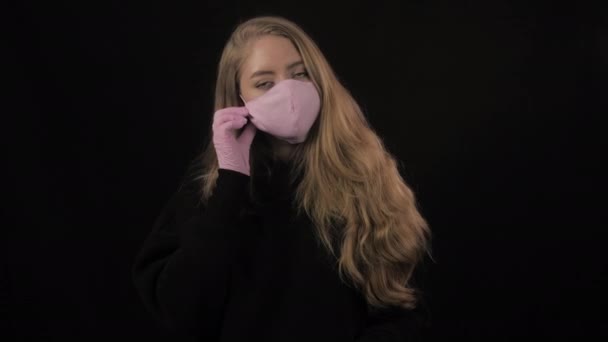 Meisje met een roze medisch masker doet het masker af en hoest. Geïsoleerd op zwarte achtergrond. Gezondheidszorg en medisch concept. Sluit het portret. 4k. Coronavirus Epidemie, ziekte, pandemie — Stockvideo