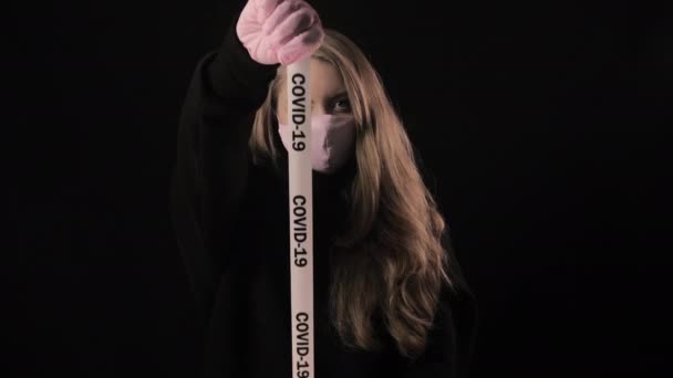 Ένα κορίτσι με ροζ μάσκα κρατάει μια κασέτα με την επιγραφή covid-19. Απομονωμένο σε μαύρο φόντο. Ιατρική περίθαλψη και ιατρική έννοια. Κοντινό πορτραίτο. 4k. Επιδημία του κερατοειδούς, ασθένεια, πανδημία — Αρχείο Βίντεο