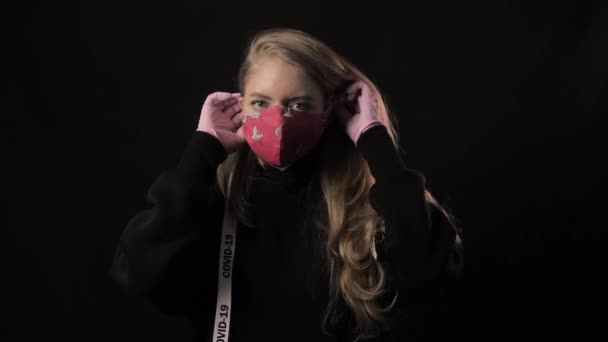 Meisje met een wit lint en de inscriptie covid 19 zet een rood leuk masker op. Geïsoleerd op zwarte achtergrond. Gezondheidszorg en medisch concept. Coronavirus Epidemisch — Stockvideo