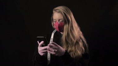 Beyaz kurdeleli ve kırmızı maskeli Covid 19 yazılı kız akıllı telefondan selfie çekiyor. Siyah arka planda izole edilmiş. Sağlık ve tıbbi konsept. Koronavirüs salgını