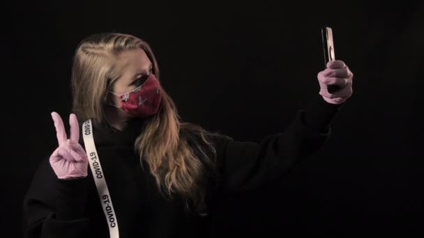 Chica con una cinta blanca y la inscripción covid 19 en máscara roja toma selfie en el teléfono inteligente. Aislado sobre fondo negro. Salud y concepto médico. Epidemia del Coronavirus — Vídeos de Stock