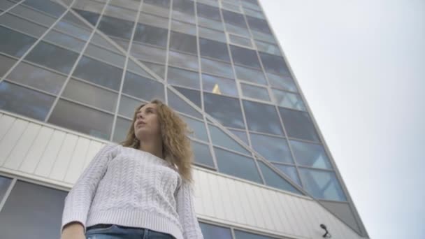 Κορίτσι σε ένα λευκό πουλόβερ με λυπημένο βλέμμα στέκεται στο παρασκήνιο ενός κτιρίου επιχειρήσεων — Αρχείο Βίντεο