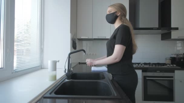 Девушка в черной одежде и маске моет руки с мылом на кухне — стоковое видео