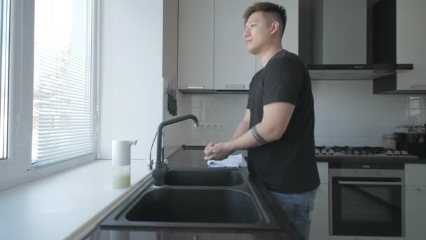 Азиатский мужчина в черной футболке и синих джинсах моет руки с мылом на кухне — стоковое видео