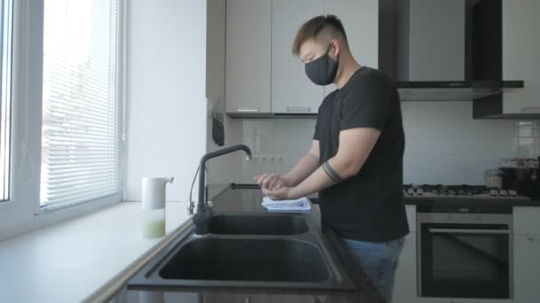 Asiatisk man med mask i svart t-shirt och blå jeans tvättar händerna med tvål i köket — Stockvideo