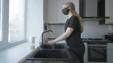 Siyah elbiseli ve maskeli bir kız mutfakta sabunla ellerini yıkıyor.