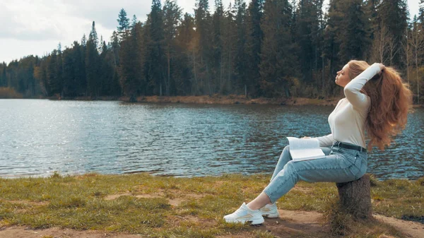 Bella giovane donna bionda seduta a leggere un libro in un parco vicino al lago. Vista laterale — Foto Stock